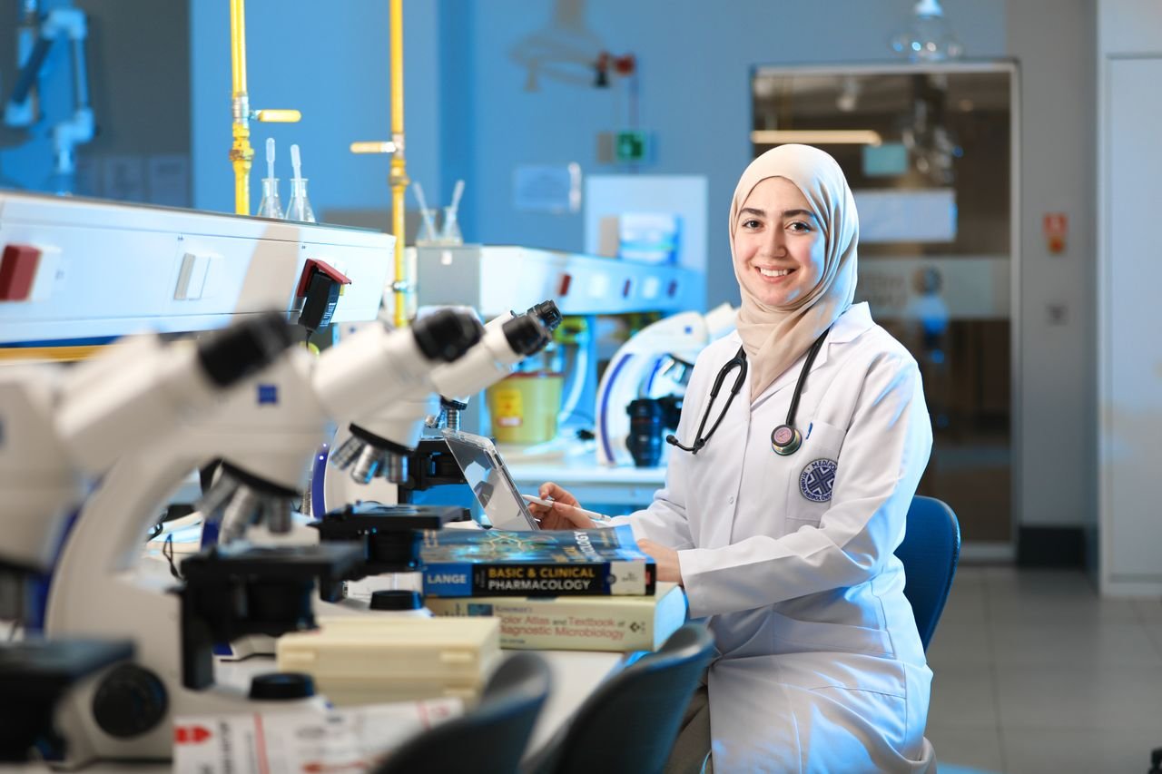 A confident female scientist in a lab coat with a hijab in a modern laboratory with microscopes and textbooks.